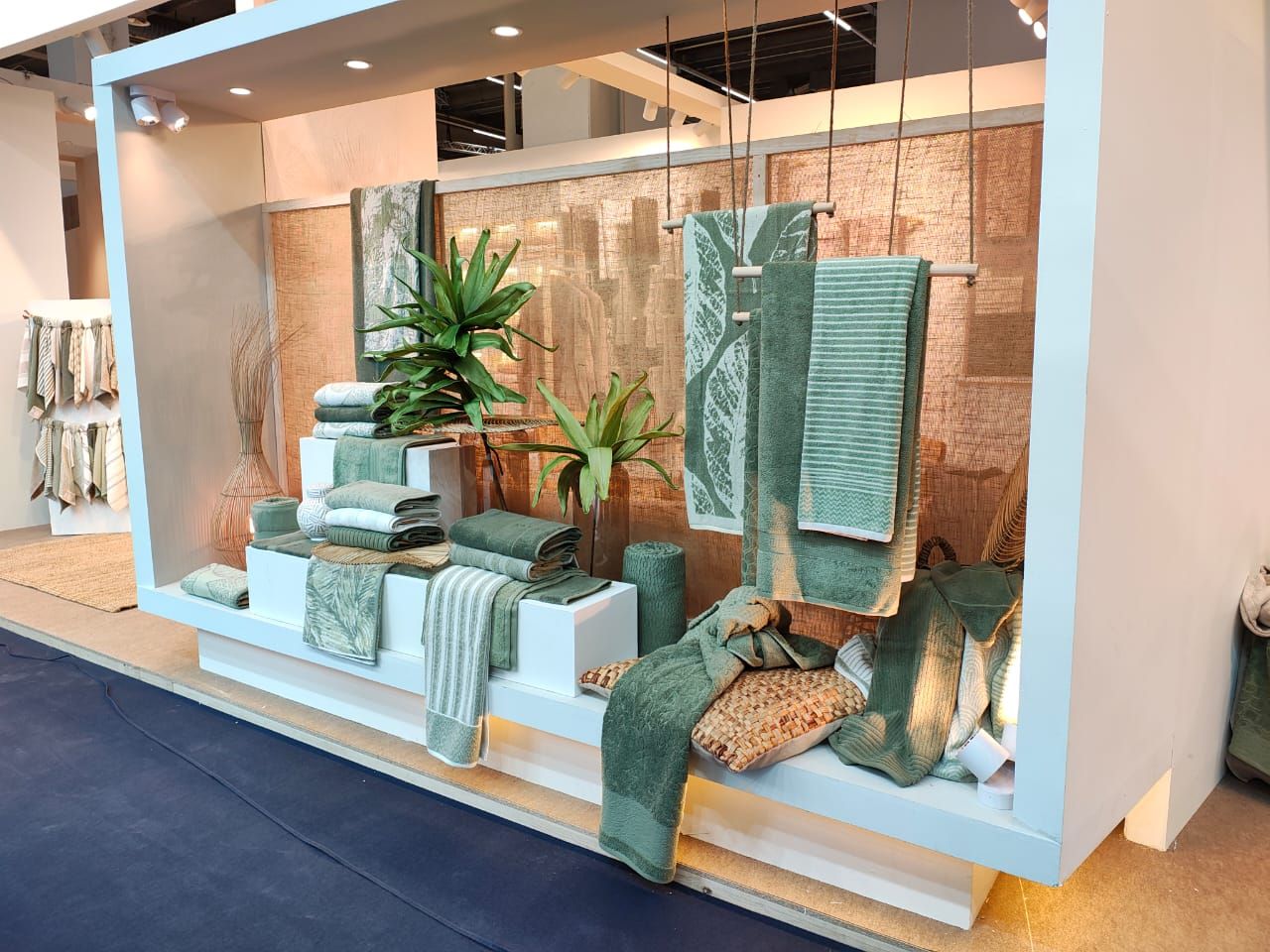 A pale blue display bay of green and teal towels, stacked and hung, with planting.